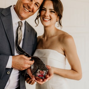 May include: A bride and groom's father smiling at the camera. The bride is holding a tie with a photo of a young boy and a man in it. The groom's father is wearing a blue suit and a patterned tie.