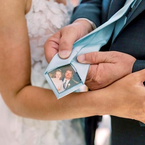 May include: A bride and groom are getting married. The groom is holding a light blue tie with a small photo of a man and a young girl tucked into the tie.