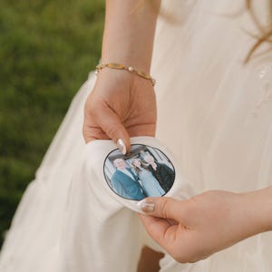 May include: A white fabric patch with a family photo is held by a person wearing a bracelet. The photo shows three people in formal attire. The background is blurred, suggesting an outdoor setting.