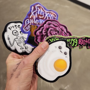 May include: A collection of four stickers featuring various designs. One sticker features a skeleton with the word "PISS" above it. Another sticker features a purple flower with the text "Kitty Bang Bang". A third sticker features a fried egg with a yolk. The fourth sticker features a mushroom with the text "Kitty Bang Bang".