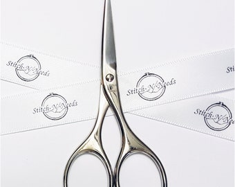 Puede incluir: Tijeras de bordado plateadas con puntas afiladas y mangos ovalados. Las tijeras están sobre una superficie blanca con una cinta blanca que tiene el texto "Stitch N/Needs" impreso.