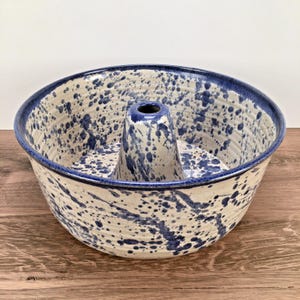 May include: A ceramic bundt pan featuring a cream-colored base with a dark blue speckled pattern throughout the interior and exterior. The round baking dish has a central chimney and a wide, flared rim with a solid dark blue painted edge.