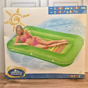 May include: A rectangular green inflatable pool float with a white perimeter rope, depicted on a product box. The box displays the text The Wet Set and Intex, with dimensions of approximately 81 1/2 inches by 52 3/4 inches printed in the corner.