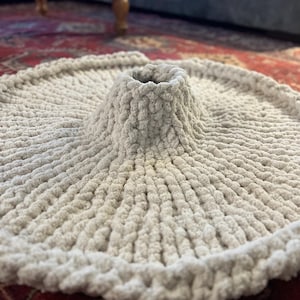 May include: A light gray, circular, knitted tree skirt with a raised center for the tree trunk. The skirt is made of a thick, textured yarn and has a soft, plush appearance. The skirt is on a red patterned rug.