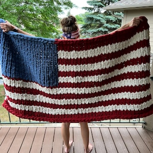 May include: A large, hand-knitted blanket in the design of the American flag. The blanket features a blue square with white stars in the upper left corner, and red and white stripes. The blanket is held up, displaying its full size and design.