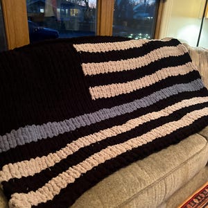 May include: A black, white, and blue crocheted blanket with a thin blue stripe across the middle, resembling a thin blue line flag.