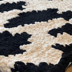 May include: A large, handmade blanket with a cow print design. The blanket features a pattern of black and cream-colored sections, creating a striking contrast. The chunky knit texture adds to its cozy appearance. This blanket is perfect for home decor.