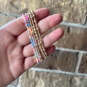 May include: A set of four stretch bracelets. Two bracelets feature small, round, metallic gold-tone beads. The other two bracelets consist of small, translucent, multi-colored beads in shades of pink, blue, orange, and green, accented with silver-tone beads.