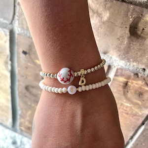 May include: Two beaded bracelets. The top bracelet is gold with a baseball-themed bead and a gold letter "D" charm. The bottom bracelet is white with a bead that has the number "8" on it. The bracelets are on a wrist.