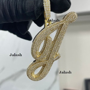 May include: A gold pendant in the shape of the letter "J" encrusted with small, clear diamonds. The pendant is held in a gloved hand.