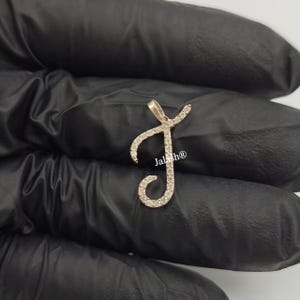 May include: A gold-toned pendant in the shape of the letter J, encrusted with small, clear gemstones. The pendant is on a black background.