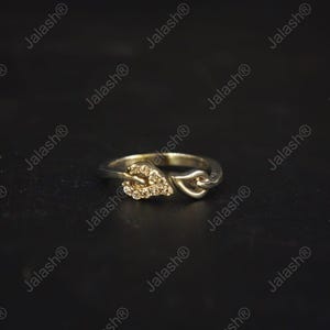 May include: A gold-colored ring with a leaf design on one side and a heart-shaped design on the other. The leaf is encrusted with small, clear gemstones. The ring is set against a dark background.
