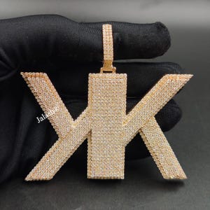 May include: Gold-toned pendant in the shape of the letters "YK" encrusted with small, sparkling stones. The pendant has a bail for a chain and is displayed against a dark background. The text "Jalash" is visible on the pendant.