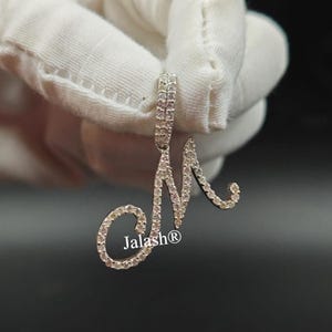 May include: A silver pendant with a cursive letter M encrusted with clear cubic zirconia stones. The pendant is on a white background.