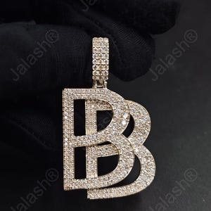 May include: A silver-toned pendant in the shape of the letter "B" with a smaller "B" intertwined. The pendant is encrusted with numerous small, sparkling stones. The bail is also covered in stones. The background is black.