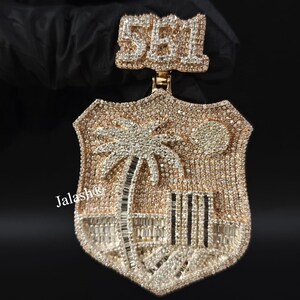 May include: A gold pendant with a shield-shaped design featuring a palm tree and the number 551 in diamonds. The pendant is covered in small diamonds.