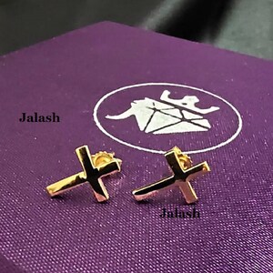 May include: A pair of gold cross earrings on a purple background. The earrings are small and simple, with a classic cross design.
