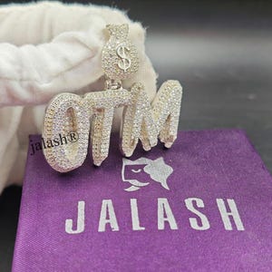 May include: Silver pendant with a money bag charm and the word "OTM" spelled out in large, bold letters with clear stones. The pendant is on a purple velvet pouch with the word "JALASH" printed on it.