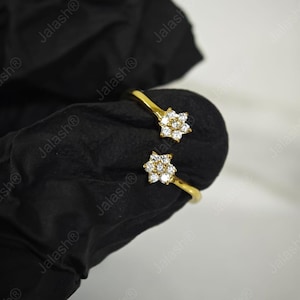May include: A gold-colored open ring featuring two flower-shaped designs. Each flower is adorned with small, clear gemstones. The ring is displayed against a black background, creating a contrast that highlights the jewelry's details.
