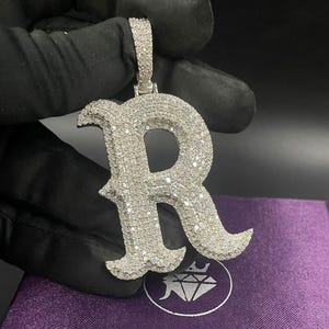 May include: A silver pendant in the shape of the letter "R", encrusted with numerous small, sparkling stones. The pendant is held by a gloved hand against a dark background, with a purple cloth visible below.