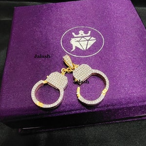 May include: A gold-tone pendant in the shape of handcuffs, encrusted with clear cubic zirconia stones. The pendant is displayed on a purple velvet box with the word "Jalash" printed on the box.