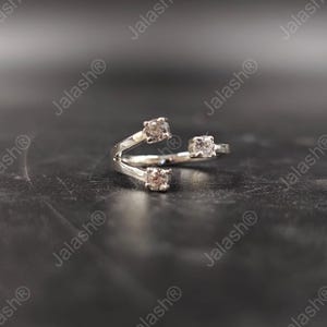 May include: A silver-toned ring with three square-cut, light-colored gemstones. The ring's design features a split shank that curves around the finger, with a gemstone at each end and one in the center. The ring is displayed on a dark surface.
