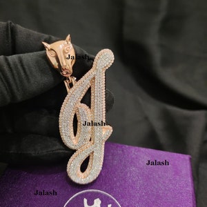 May include: A rose gold pendant featuring the letter 'J' encrusted with clear and white cubic zirconia stones. The pendant is attached to a small rose gold lion head charm. The pendant is displayed on a purple velvet box with the brand name "Jalsh" printed on it.