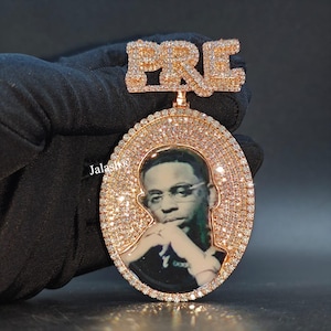 May include: A rose gold pendant with a photo of a man wearing glasses, surrounded by a halo of small, sparkling stones. The word "PRE" is spelled out in large, gem-studded letters above the oval pendant. The pendant is held by a gloved hand.
