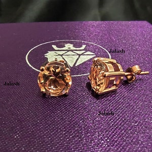 May include: A pair of rose gold earrings with a large, round, clear gemstone in the center of each earring. The earrings are set on a purple background with the word "Jalash" printed on the background.