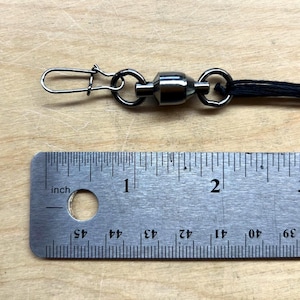 May include: A dark metallic fishing swivel with a snap connector attached to a black cord, positioned above a metal ruler that shows the swivel assembly measures approximately 1.5 inches in total length from the snap tip to the cord attachment.