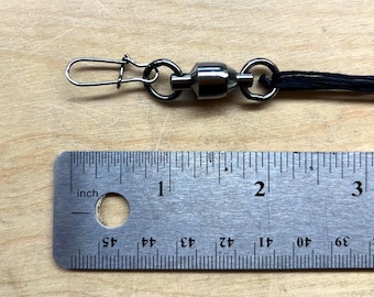 May include: A dark metallic fishing swivel with a snap connector attached to a black cord, positioned above a metal ruler that shows the swivel assembly measures approximately 3.8 cm in total length from the snap tip to the cord attachment.