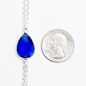 K&ouml;nnte beinhalten: Eine silberne Kettenkette mit einem tropfenf&ouml;rmigen blauen Edelstein-Anh&auml;nger. Der Anh&auml;nger ist etwa so gro&szlig; wie eine US-amerikanische 25-Cent-M&uuml;nze.