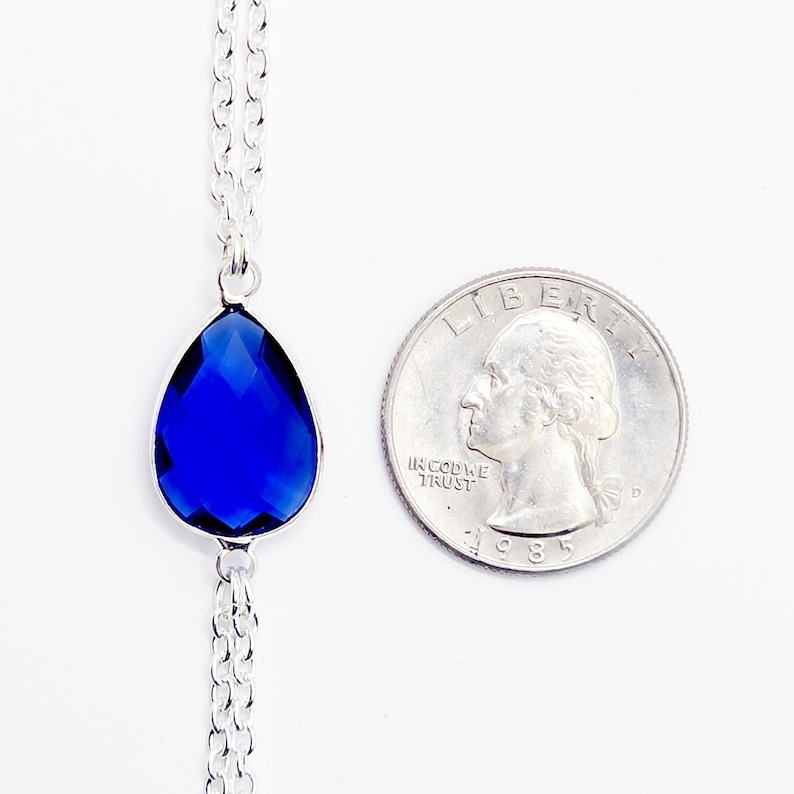 K&ouml;nnte beinhalten: Eine silberne Kettenkette mit einem tropfenf&ouml;rmigen blauen Edelstein-Anh&auml;nger. Der Anh&auml;nger ist etwa so gro&szlig; wie eine US-amerikanische 25-Cent-M&uuml;nze.