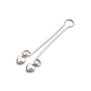 May include: A pair of silver-colored nipple clamps. Each clamp features a long, slender bar with a loop at one end and a small, heart-shaped charm dangling from the other end. The clamps are designed to be worn on the nipples.
