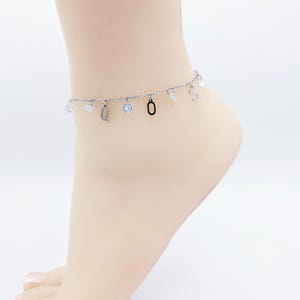May include: A silver anklet with dangling charms, including clear crystals, the letter "Q", the number "0", and the letter "S". The anklet is worn on a foot, showcasing its delicate design and sparkling accents.