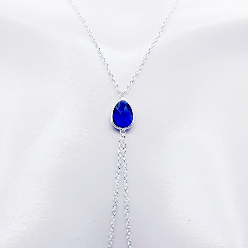 K&ouml;nnte beinhalten: Eine silberne Kettenkette mit einem tropfenf&ouml;rmigen blauen Edelstein-Anh&auml;nger. Die Kette ist unten verdoppelt und bildet zwei Str&auml;nge.