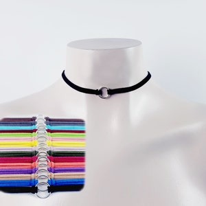 May include: A black suede choker necklace with a silver ring accent. The necklace is displayed on a mannequin. A variety of colors of the same choker are shown in the background.