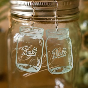 May include: Clear acrylic earrings shaped like Ball mason jars, with the word "Ball" in a script font. The earrings hang from silver-colored fish hook ear wires. The background shows a mason jar.