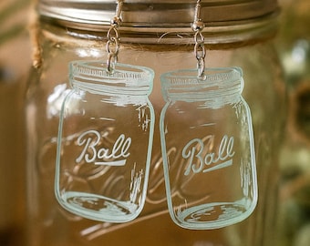 May include: Clear acrylic earrings shaped like Ball mason jars, with the word "Ball" in a script font. The earrings hang from silver-colored fish hook ear wires. The background shows a mason jar.