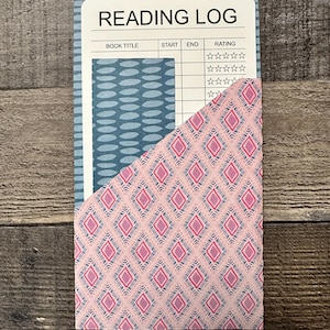 May include: A Reading Log notebook with a blue and white oval pattern on the left side and a pink diamond pattern pocket. The notebook has a white top section with the words "READING LOG" and spaces for book title, start, end, and rating.