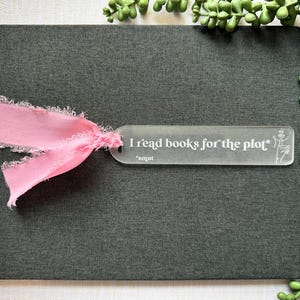 May include: A dark gray book with a pink ribbon bookmark. The bookmark is clear with the text "I read books for the plot" and a small illustration. Green plant decorates the background.