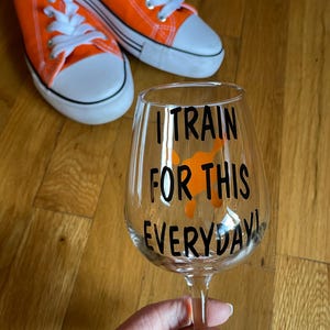 May include: A clear glass wine glass with the text "I train for this everyday!" in black lettering. The glass is decorated with an orange silhouette of a person running.