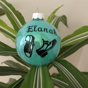 May include: A teal blue Christmas ornament with glitter and black silhouettes of shoes. The ornament is personalized with the name "Elanah".