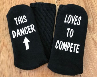 May include: A pair of black socks with white text. One sock says "This Dancer" with an arrow pointing up. The other sock says "Loves to Compete".