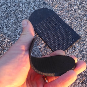 Könnte beinhalten: Zwei schwarze, flexible, halbkreisförmige Einsätze mit einer texturierten, gerippten Oberfläche. Ein Teil wird in der Hand gehalten und zeigt seine Biegsamkeit, während das andere flach liegt. Diese Komponenten haben eine abgerundete Kante.