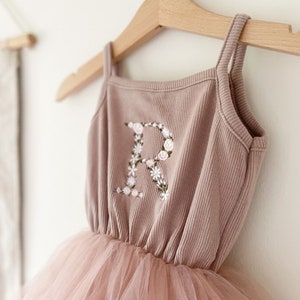 Op de afbeelding: Een stoffig roze tule tutu-jurk met een bijpassend geribd tanktop. Het tanktop heeft een met bloemen geborduurde letter "R" aan de voorkant.