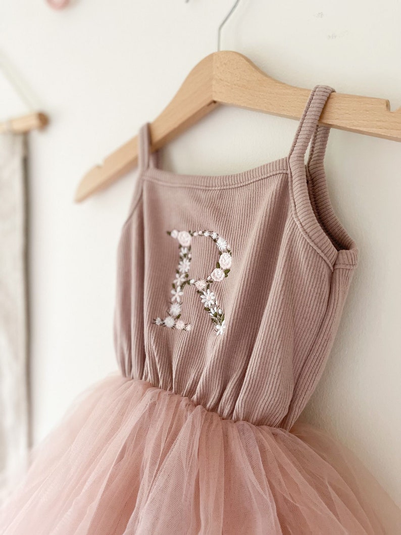 Op de afbeelding: Een stoffig roze tule tutu-jurk met een bijpassend geribd tanktop. Het tanktop heeft een met bloemen geborduurde letter "R" aan de voorkant.