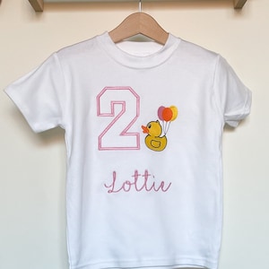 May include: White t-shirt with pink embroidered number 2, a yellow rubber duck with pink and orange balloons, and the name "Lottie" underneath.