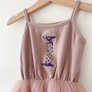 Op de afbeelding: Een roze tule tutu-jurk met een roze geribbeld tanktop met een geborduurde bloemletter I.