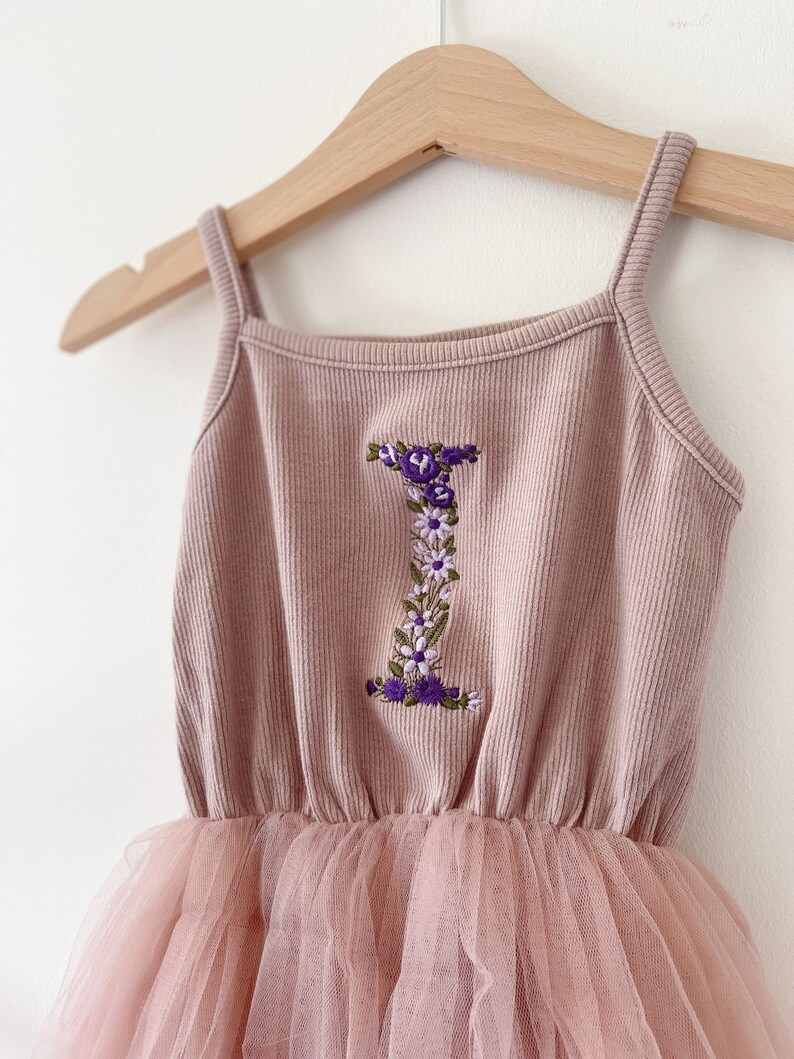 Op de afbeelding: Een roze tule tutu-jurk met een roze geribbeld tanktop met een geborduurde bloemletter I.